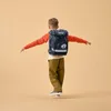 A young boy wearing a backpack is balancing with his arms outstretched against a plain background. Produktbild-Vorschau