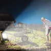 A person is digging in a garden at night, illuminated by a work light. A wheelbarrow is nearby. Voir les images du produit