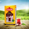 A package of Caykur Rize tourist tea is displayed in front of a lush green tea plantation under a bright sunny sky. A glass of prepared Turkish tea with sugar cubes is also visible on a wooden surface. Anteprima dell’immagine prodotto