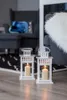 Two white lanterns with lit candles are placed on a wooden floor in front of a gray armchair and a black and white patterned rug. Anteprima dell’immagine prodotto
