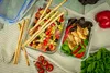 Two glass food containers filled with salads and chicken, along with breadsticks and tomatoes, are arranged on a checkered cloth. Produktbild-Vorschau