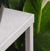 A close-up view of a silver plant stand with a blurred green plant in the background. Anteprima dell’immagine prodotto
