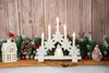 A Christmas-themed decoration featuring a white candle holder with snowflake designs, LED candles, a small illuminated tree, and surrounding festive elements like pine cones, greenery, and a small house. Náhled obrázku produktu