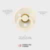A close-up, top-down view of a cream-colored pacifier with a gold ring around the nipple, set against a light gray background. Text overlays highlight product features and origin. Produktbild-Vorschau