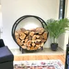 A round black metal firewood rack filled with logs is placed next to a potted palm plant in a living room setting. Náhľad obrázku produktu