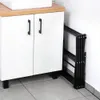 A white bathroom cabinet with black handles and a folded black drying rack leaning against the wall next to it. Produktbild-Vorschau