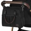 A black diaper bag is attached to the handle of a stroller. Produktbild-Vorschau