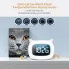 A cute cat ear alarm clock is displayed on a shelf next to a book titled 'The Cat Book'. Produktbild-Vorschau
