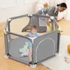 A baby is playing in a playpen while a person in the background cleans. Podgląd zdjęcia produktu