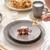 A table setting with a slice of cake on a plate, a plate of pasta in the background, and a mug of milk. Náhled obrázku produktu