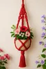 A red macrame plant hanger with a beige pot filled with pink and red flowers and greenery, hanging against a light gray wall. Purple bellflowers are visible on the right side of the image. Produktbild-Vorschau