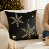 A black pillow with gold snowflakes is placed on a white couch next to a person wearing a beige sweater. A Christmas tree with lights is visible in the background. Voir les images du produit