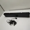 A black soundbar and its power adapter are placed on a white surface against a white wall. Produktbild-Vorschau