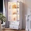 A white display cabinet with glass doors and drawers, illuminated by LED lights, stands in a room next to a window with curtains and a fireplace. Produktbild-Vorschau