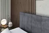 A close-up view of a bed with a dark gray upholstered headboard, a bedside table with a lamp and a vase, and a slatted wooden wall in the background. Produktbild-Vorschau