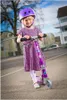 A young girl wearing a purple helmet and dress is standing on a scooter on an outdoor paved area. Produktbild-Vorschau