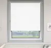 A white roller blind is installed on a window, covering most of the glass. The window frame is white, and the wall around it is light gray. A small framed picture is visible on the right wall. Produktbild-Vorschau