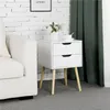 A white nightstand with two drawers sits next to a white sofa, with a white brick wall and green curtains in the background. Produktbild-Vorschau