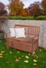 A wooden storage bench with two white cushions sits on a grassy lawn in front of a stone wall, with autumn trees in the background. Produktbild-Vorschau