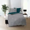 A bed with bedding is set up in a room with a plant and a bedside table with a lamp.