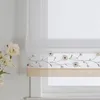A close-up view of a window with a roman blind featuring a floral embroidery pattern. A bouquet of white flowers is visible on the left side of the image. Produktbild-Vorschau
