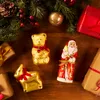 Christmas-themed chocolate figures and gifts arranged on a wooden surface with festive greenery. Náhled obrázku produktu