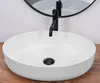 A white basin sink with a black faucet and drain is shown on a wooden surface, with a round mirror in the background. Produktbild-Vorschau
