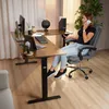 A woman is sitting at a height-adjustable L-shaped desk, talking on the phone. The desk has a monitor, laptop, keyboard, and microphone on it. Produktbild-Vorschau