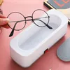 A hand is holding a pair of round glasses over a white ultrasonic cleaner. Produktbild-Vorschau