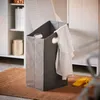 A laundry basket with clothes hanging on it, placed on a rug next to a cabinet and a shelf with jars. Náhled obrázku produktu