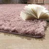 A close-up view of a fluffy pink rug with an open book on it, placed on a wooden floor. Produktbild-Vorschau