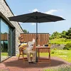 An elderly man sits on a chair reading a newspaper under a large black umbrella on a wooden deck. A small table with a cup and books is nearby. The background shows a garden with lush greenery and a stone wall. Voir les images du produit