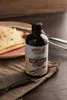 A bottle of vanilla extract is placed on a brown cloth next to a plate of crepes and vanilla beans. Produktbild-Vorschau