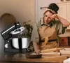 A woman is tiredly wiping her forehead while baking in a kitchen, with a stand mixer and baking ingredients on the counter. Produktbild-Vorschau