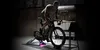 A man is riding a stationary bicycle in a dimly lit garage or workshop. Produktbild-Vorschau