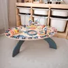 A wooden activity table for children is shown in a room with storage bins in the background. Podgląd zdjęcia produktu