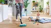A woman is vacuuming a wooden floor while a baby sits on a playmat nearby. Produktbild-Vorschau
