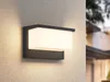 A modern outdoor wall light mounted on a textured wall, with a blurred house in the background. Podgląd zdjęcia produktu