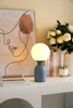 A blue table lamp with a white globe shade is placed on a white shelf next to a framed abstract art piece and a vase of white flowers. Books and a candle are also on the shelf. Podgląd zdjęcia produktu
