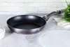 A frying pan is placed on a countertop next to a small white bowl and a green plant. Podgląd zdjęcia produktu