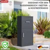 A dark gray parcel box with a letter slot and a door, placed outdoors in front of a house with plants and stairs. Náhled obrázku produktu