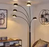 A modern living room corner with a multi-headed floor lamp, a framed abstract art piece, a shelf with decorative items, and a part of a sofa. Produktbild-Vorschau