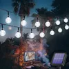 An outdoor patio scene at dusk with string lights, a television playing, and a fire pit. Anteprima dell’immagine prodotto