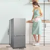 A woman in an apron stands next to a refrigerator in a kitchen setting. Produktbild-Vorschau