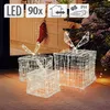 Three illuminated gift boxes with bows, set up outdoors near a Christmas tree and a house facade. Produktbild-Vorschau