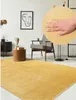 A living room scene with a yellow rug, a chair, a side table, and artwork on the wall. A close-up of the rug texture is shown in a circle. Produktbild-Vorschau
