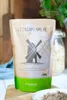A bag of flaxseed flour on a wooden surface, with a jar of oats and greenery in the background. Produktbild-Vorschau