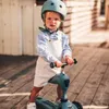 A young child wearing a helmet and overalls stands on a wooden deck, holding the handlebars of a blue scooter. Podgląd zdjęcia produktu