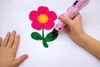 A child's hands are using a pink 3D pen to draw a flower on white paper. Another child's hand is visible on the left side of the image. Náhľad obrázku produktu