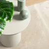 A close-up view of a beige rug with a small round table on top, featuring a green plant and two green candles. Produktbild-Vorschau